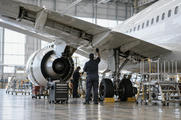 Synthetic editorial illustration. An anonymous aircraft maintenance hangar with a realistic unmarked narrow-body passenger aircraft and small mechanics seen from behind beneath the wing, industrial scale and diffuse daylight.