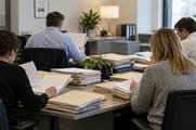 Synthetic editorial illustration. An ordinary commercial bank back-office credit team reviewing paper folders around a desk, anonymous people seen from behind, credible office interior and no readable forms or computer displays.