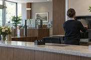 Synthetic editorial illustration. An anonymous bank branch interior at opening time with a staff member seen from behind organizing plain closed folders behind a modest counter, reassuring ordinary architecture, no coins or miniature vault.