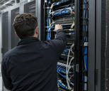 Synthetic editorial illustration. An anonymous information-security technician seen from behind inspecting physical network connections in a restrained operational server room, real cabling and secure cabinet hardware, no crisis or hacker symbolism.