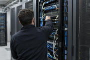 Synthetic editorial illustration. An anonymous information-security technician seen from behind inspecting physical network connections in a restrained operational server room, real cabling and secure cabinet hardware, no crisis or hacker symbolism.
