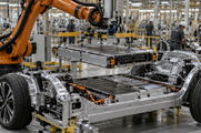 Synthetic editorial illustration. A practical electric-vehicle assembly station with unbranded vehicle chassis and credible battery modules being installed by robotic equipment, small workers in the background, no branding.
