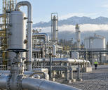 Synthetic editorial illustration. A working natural-gas processing plant with credible pipes, separators and storage vessels, a distant maintenance worker for scale, cool morning daylight, no smoke spectacle.