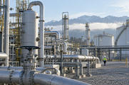 Synthetic editorial illustration. A working natural-gas processing plant with credible pipes, separators and storage vessels, a distant maintenance worker for scale, cool morning daylight, no smoke spectacle.
