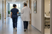 Synthetic editorial illustration. A quiet modern outpatient-care facility with two anonymous healthcare staff seen from behind walking down a functional corridor, practical consultation rooms and natural window light, no signs or patient faces.