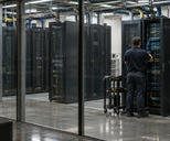 Synthetic editorial illustration. An anonymous secure institutional data facility with realistic metal server cabinets behind a glass partition and a small maintenance technician seen from behind, practical cabling and calm lighting.