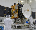 Synthetic editorial illustration. A generic communications satellite with believable antenna and solar-array assemblies undergoing integration in an aerospace cleanroom, small technicians seen from behind for scale.