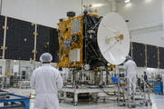 Synthetic editorial illustration. A generic communications satellite with believable antenna and solar-array assemblies undergoing integration in an aerospace cleanroom, small technicians seen from behind for scale.