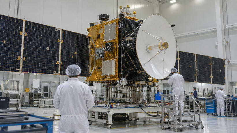 Synthetic editorial illustration. A generic communications satellite with believable antenna and solar-array assemblies undergoing integration in an aerospace cleanroom, small technicians seen from behind for scale.