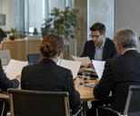 Synthetic editorial illustration. An anonymous corporate investor-relations meeting in a modest glass office, several colleagues seen obliquely from behind reviewing blank-backed report pages, real office depth and no visible monitors or charts.