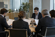 Synthetic editorial illustration. An anonymous corporate investor-relations meeting in a modest glass office, several colleagues seen obliquely from behind reviewing blank-backed report pages, real office depth and no visible monitors or charts.