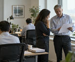 Synthetic editorial illustration. Anonymous financial analysts in a modest open-plan research office, a senior colleague discussing blank-backed paper reports beside a window, other workstations turned away, calm natural light.