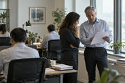 Synthetic editorial illustration. Anonymous financial analysts in a modest open-plan research office, a senior colleague discussing blank-backed paper reports beside a window, other workstations turned away, calm natural light.