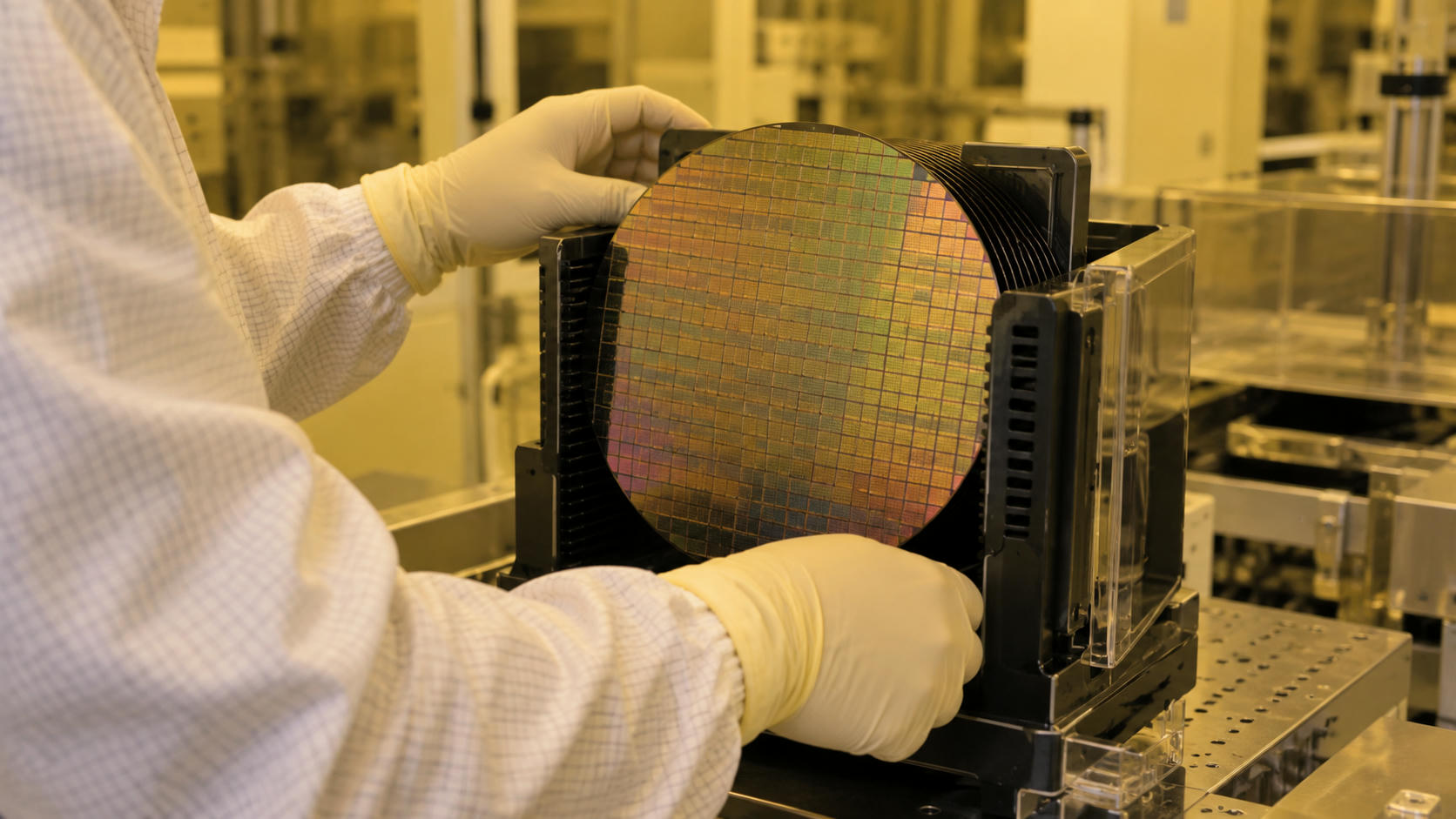Synthetic editorial illustration. A real-scale silicon wafer under neutral amber cleanroom light in an anonymous semiconductor fabrication facility, with a gloved technician's hands operating a credible wafer carrier; no exaggerated glowing chips.