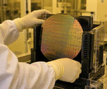 Synthetic editorial illustration. A real-scale silicon wafer under neutral amber cleanroom light in an anonymous semiconductor fabrication facility, with a gloved technician's hands operating a credible wafer carrier; no exaggerated glowing chips.