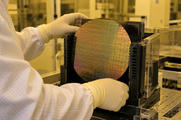 Synthetic editorial illustration. A real-scale silicon wafer under neutral amber cleanroom light in an anonymous semiconductor fabrication facility, with a gloved technician's hands operating a credible wafer carrier; no exaggerated glowing chips.